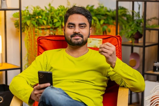Indian man use credit bank card and smartphone while transferring money, purchases online shopping cashless buy order food delivery at home apartment indoors. Smiling Arabian guy sits in room on chair