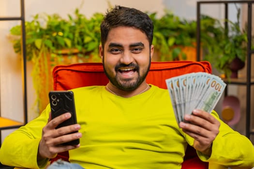 Planning budget. Rich excited Indian man use smartphone calculator app count money cash calculate domestic bills salary at home room. Guy satisfied of income earnings win saves money for planned gifts