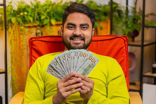 Successful happy rich business Indian man counting money cheering up with high profits, celebrate lottery game win, satisfied of wealth income earnings salary, holding dollars cash in hands at home