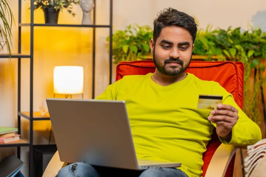 Indian man using credit bank plastic card and laptop computer while transferring money, purchases online shopping, order food delivery on internet at modern home room apartment. Guy in sits on chair