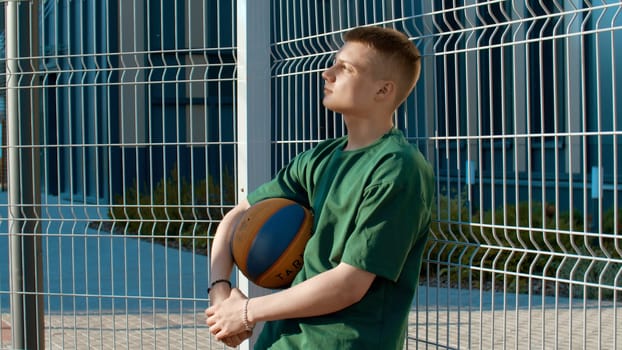 Young sportsman leaning on fence and holding basketball ball. Media. Portrait of fit caucasian man posing on sports ground in urban city outdoors