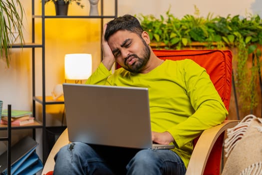 Displeased sad Indian Arab man use laptop notebook typing browsing working, loses becoming surprised sudden lottery results bad news fortune loss game fail computer virus. Arabian guy at home on chair