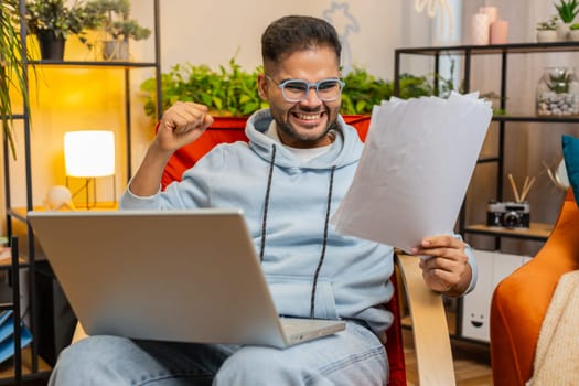 Stressed Indian man working on laptop at home room looking at unpaid bank debt bills doing paperwork planning budget calculate finances mortgage payments. Angry displeased guy scream on paper bills