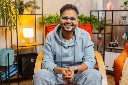 Portrait of happy calm Indian man at home chair smiling friendly, glad expression looking away dreaming resting, relaxation feel satisfied good news, celebrate win. Arabian young guy in living room