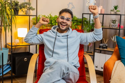 Happy Indian man shouting, celebrating success, winning, goal achievement good news lottery luck victory, watching TV championship at modern home apartment. Arabian guy in living room sits on chair