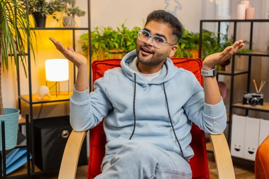 What. Why. Frustrated Indian man raising hands in indignant expression, asking reason of failure, demonstrating disbelief irritation by troubles, quarrel at home room apartment. Guy sits on chair