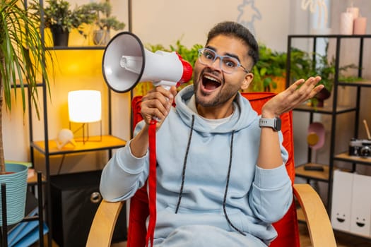 Excited Indian man talking with megaphone, proclaiming news, loudly announcing advertisement promotion sale message, warning using loudspeaker to shout speech at home room apartment. Guy sits on chair