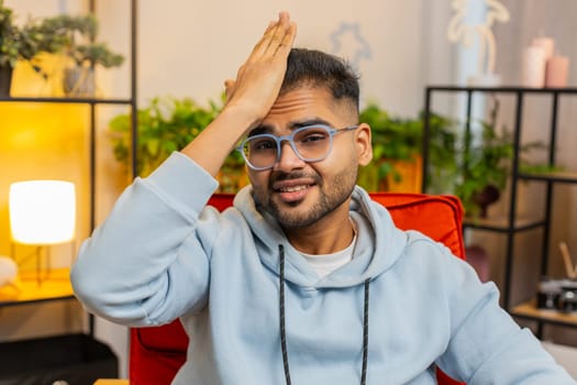 Face palm. Shame on you. Upset Indian man making disrespect gesture, feeling bored, disappointed in fail results, bad news. Unhappy confused Arabian guy at home apartment living room sitting on chair