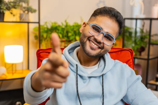 Hey you. Happy satisfied Indian man smiling excitedly and pointing to camera, choosing lucky winner, indicating to awesome you, inviting approve. Arabian Hindu guy at home living room sitting on chair