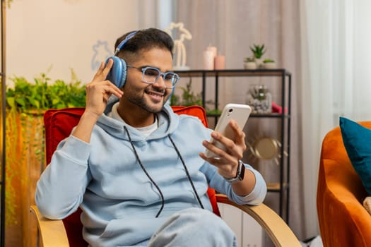 Happy relaxed overjoyed Indian Arabian man in wireless headphones dancing on chair at home choosing listening favorite energetic disco pop music in smartphone entertaining. People weekend activities
