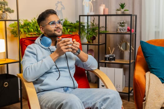 Attractive smiling Indian man drinking a cup of warm coffee or herbal tea sitting at home living room rocking chair in morning. Arabian guy enjoying comfortable relaxing alone resting, taking a break