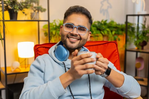 Attractive smiling Indian man drinking a cup of warm coffee or herbal tea sitting at home living room rocking chair in morning. Arabian guy enjoying comfortable relaxing alone relaxing, taking a break