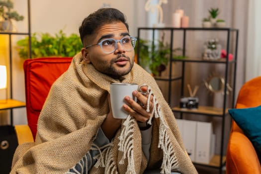 Sick Indian man in plaid sit alone shivering from cold on chair drinking hot tea in unheated apartment without heating due debt. Unhealthy Arabian guy sit on chair feeling discomfort try to warming up