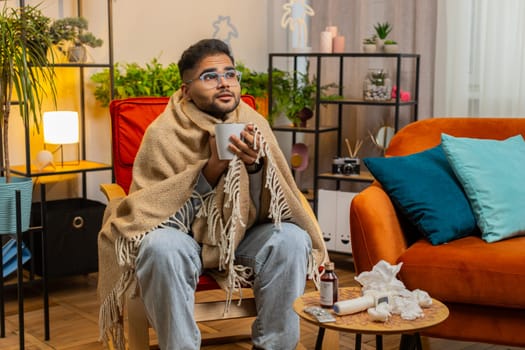 Sick Indian man in plaid sit alone shivering from cold on chair drinking hot tea in unheated apartment without heating due debt. Unhealthy Arabian guy sit on chair feeling discomfort try to warming up