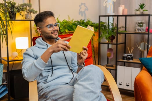 Indian man reading interesting book, turning pages smiling enjoying literature, taking a rest on comfortable chair. Portrait of peaceful cheerful guy relaxing at home apartment living room on chair