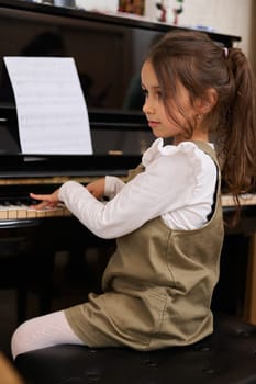 Confident portrait of an inspired beautiful talented little kid girl learning music, performing the rhythm of classical music while playing piano, putting fingers on keys and dreamily looking away