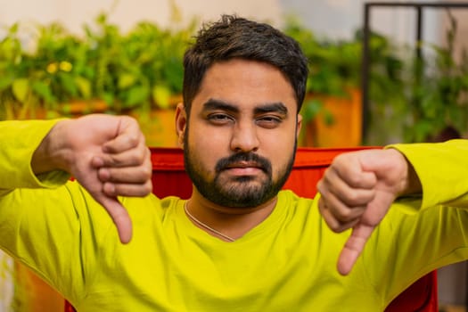 Dislike. Portrait of upset unhappy Arabian man showing thumbs down sign gesture, expressing discontent, disapproval dissatisfied bad work at home apartment indoors. Displeased Indian guy sits on chair