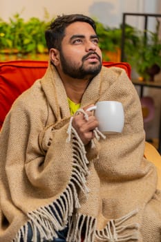 Sick Indian man in plaid sit alone shivering from cold on sofa drinking hot tea in unheated apartment without heating due debt. Unhealthy Arabian guy sits on chair feeling discomfort try to warming up