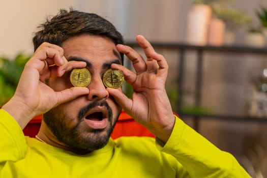 Indian man freelancer holding two gold coins, successful developer programmer. Guy stock trader earning bitcoins after online monitoring trading operations. Increasing wealth, financial prosperity