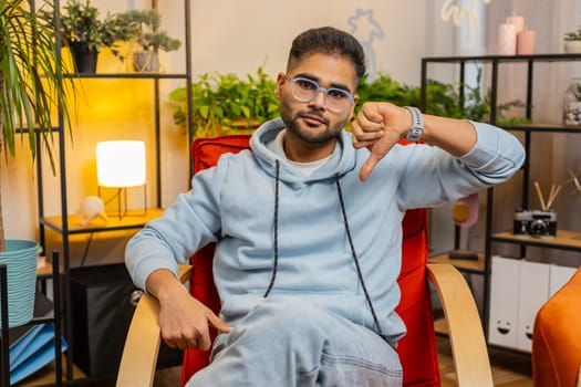 Dislike. Upset unhappy young Indian man showing thumbs down sign gesture, expressing discontent, disapproval dissatisfied bad work at home apartment indoors. Displeased Arabian guy in room on chair