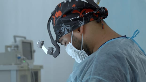 A close up male medical worker with a disposable mask and hat is wearing a special equipment on his head. Action. Surgeon at work in modern clinic