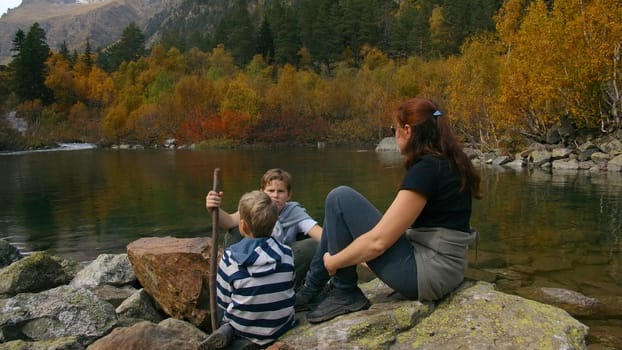 Family is sitting on rocks by river. Creative. Family vacation in nature by river in autumn forest. Mother and children are resting on rocks by water in mountain forest in autumn.