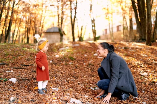 Little girl stands in front of her mother squatting in the autumn forest touching the fallen leaves. High quality photo