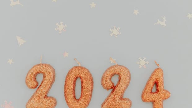 Shiny light brown candles number 2024 lie with white blurred shiny festive confetti lie below on a pastel blue background with copy space on top, flat lay close-up.