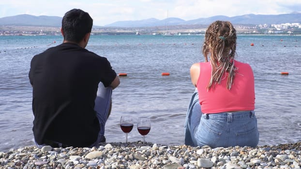 Rear view of a couple sitting at seaside with red wine glasses standing near on a summer sunny day. Man in black shirt and woman with afro braids enjoying marine landscape.