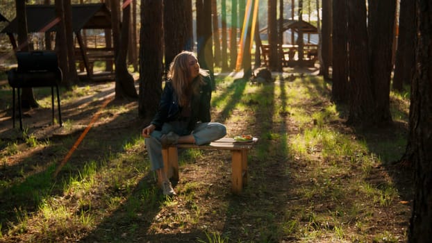 Girl sitting on picnic bench in green park near camp among trees. Stock footage. Woman spending time alone outdoors