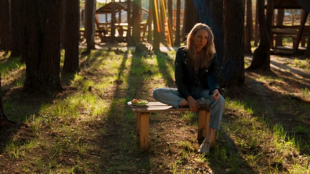 Pensive blond girl sitting alone on a small bench with a plate of vegetables. Stock footage. Woman in outdoors camp in the forest