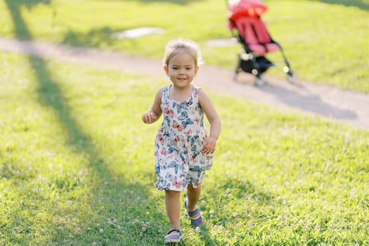 Little girl walks smiling along the green lawn from the stroller on the path. High quality photo