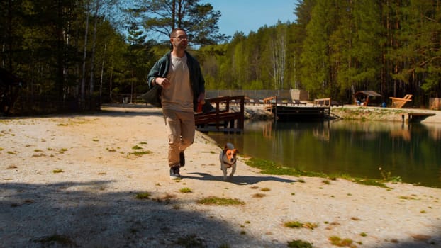 Happy man and his little cute puppy running along the green small lake. Stock footage. Man jogging with jack russell