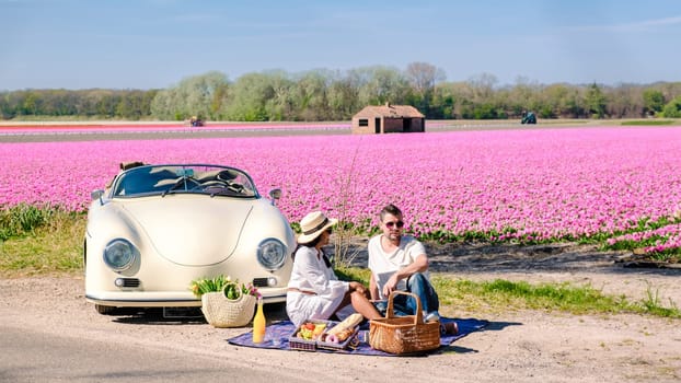 couple on a roadtrip in the Netherlands with an old vintage car, men, and woman having a picnic with on the background a pink flower field during spring