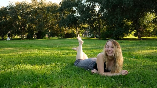Happy carefree woman lying on green grass outdoors in a city park. Concept. A young beautiful girl with blonde curly hair enjoying a nice sunny day and laughing