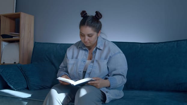Young cute woman reading book indoors. Media. Beautiful plus size girl at home sitting on couch with a book