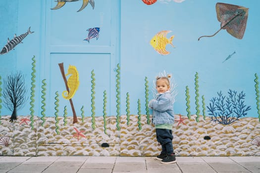 Little girl stands leaning against a wall with colorful drawings. High quality photo