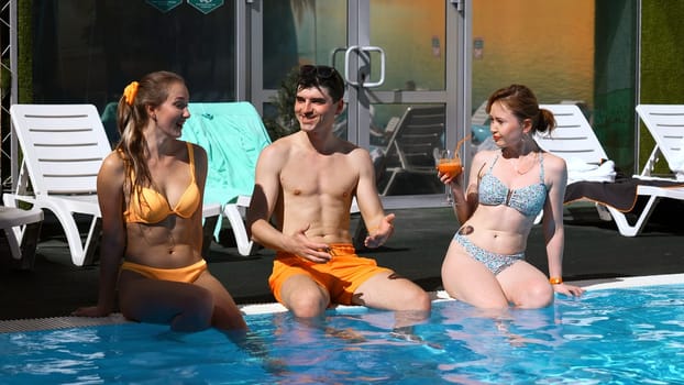 Three friends talking and having fun on the edge of the outdoors swimming pool. Clip. Two women and a man relaxing by the hotel pool