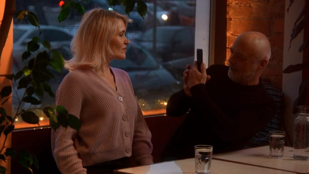 Elegant senior woman is posing for smartphone camera during dinner with husband in cafe. Stock footage. Modern devices and photography concept