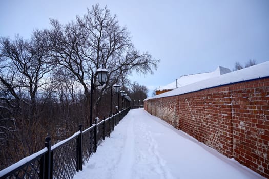 Snow-covered road in a winter park next to an old brick wall