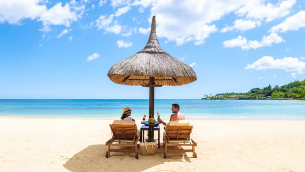 Man and Woman on a Tropical Beach with Chairs And Umbrella on a Tropical Beach. Beautiful tranquil white sand beach with two chairs and thatched umbrella, couple drinking cocktails on the beach
