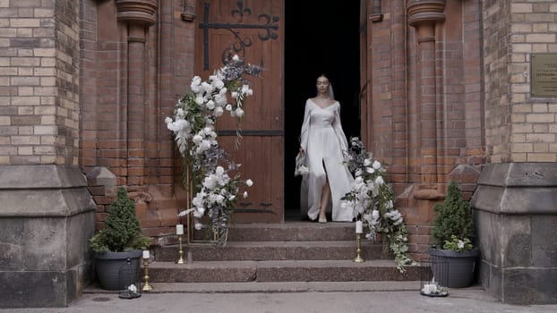 A young bride coming out of their temple. Action.A beautiful girl in a white dress with flowers who comes out of a dark building and there is an arch of white flowers nearby. High quality 4k footage