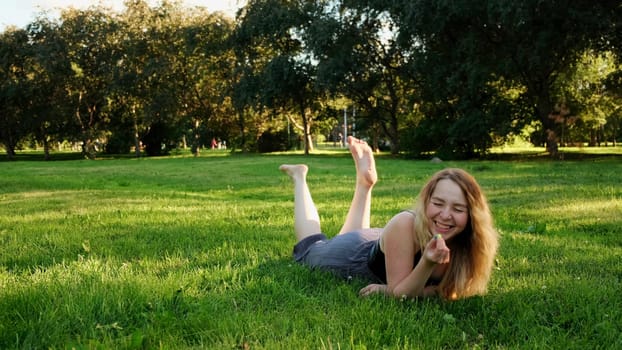 A cheerful woman in a clearing. Concept. A beautiful woman with long curly hair lying on the grass swinging her legs and laughing. High quality 4k footage