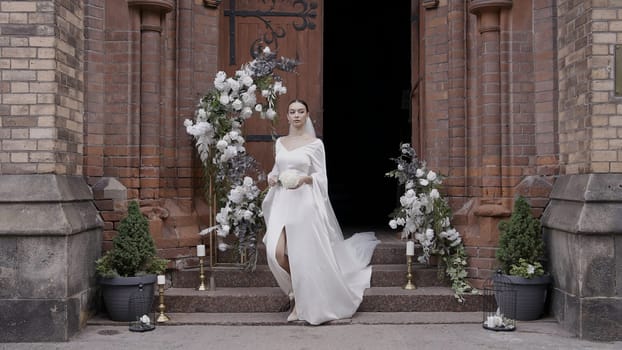A young bride coming out of their temple. Action.A beautiful girl in a white dress with flowers who comes out of a dark building and there is an arch of white flowers nearby. High quality 4k footage