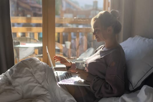 Happy casual beautiful woman working on a laptop sitting on the bed in the house