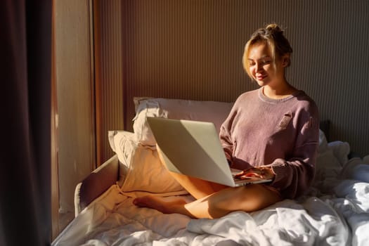 Happy casual beautiful woman working on a laptop sitting on the bed in the house