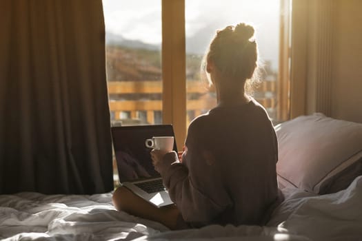 Happy casual beautiful woman working on a laptop sitting on the bed in the house