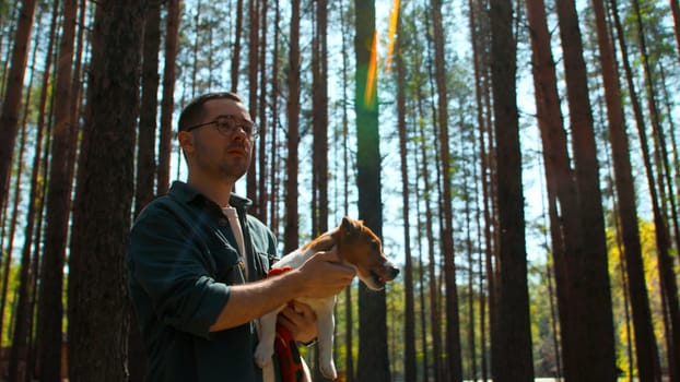 Pensive man in glasses standing among trees in forest with a dog in his hands. Stock footage. Young man in shirt and his puppy