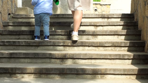 Close up of woman helping her little child boy to climb the stairs by holding his hand. Mother with small kid son walking up the stairs.