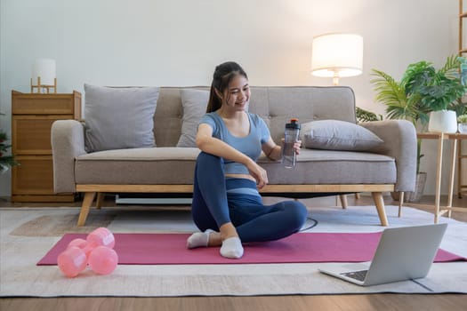 Young Asian woman using laptop and drinking water while doing yoga exercise at home. relax time.